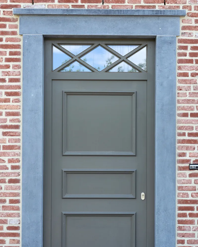 Porte d'entrée pleine gris vert avec imposte vitrée, encadrement en pierre bleue dans un mur de brique